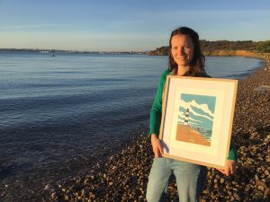 Noémie Courant stands on a beach with one of her linocuts depicting a lighthouse