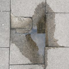 A puddle of water on a pavement. The paving slabs are cracked.