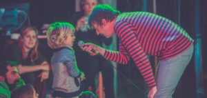 An adult performer (artist Kid Carpet) bending over and holding a microphone for a toddler to speak into. Audience seated on the floor are looking on. 