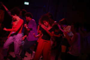 A group of performers captured mid-movement with arms raised and bathed in a red light. 