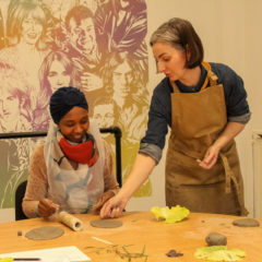 Rebecca May stands in an apron, guiding a seated workshop participant.