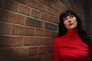 Image Credit: Michelle Adamson Louise Fazackerley stands against a brick wall. Her hair is dark with a fring and she wears a red turtleneck top and red lipstick.