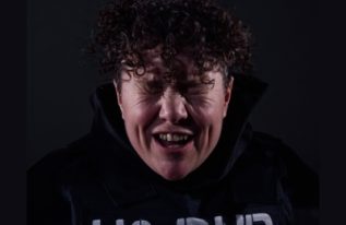 Jess Thom, a white woman in her early forties with short curly brown hair and dimples. She is wearing a black hoodie with a black stab vest over it. The stab vest has the words ‘NO DNR’ stencilled onto it in white paint. She has her closed and scrunched and has an expression of joy.