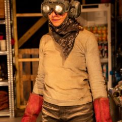 A woman is pictured with large earphones over their ears, they are also wearing goggles over their eyes. This person has a scarf wrapped around their neck and is wearing red rubber gloves. They are wearing a grey jumper and paint splattered jeans. They are pictured in a bunker setting.