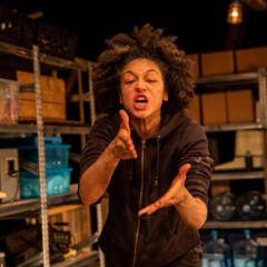 Woman stairs directly into the camera with her arms in front of her, palms facing inward. The picture of woman has captured her mid sentence, her mouth is slightly open as if she is saying something powerful, with meaning. The woman is wearing a dark hoodie with her sleeves rolled up. Behind the woman are metal shelving, like you would find in a bunker.