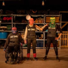 A behind the scenes image, Three people stare directly at the camera and are each wearing different coloured, knitted balaclavas where only their eyes and mouths are exposed. They are all wearing black stab vests with the words ‘NO DNR’ stencilled onto the vests in white paint. Jess is on the left is in a black wheelchair holding the hand of Chopin, who is stood in the centre of the image. She is wearing a red and orange knitted balaclava with her left arm rested upon the shoulder of Charmaine who is standing on the right of the image. She is wearing a knitted green balaclava with her hand on Chopin's shoulder.