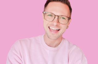 Image of Nathaniel, a white man with short brown hair, glasses and wearing a pink jumper. He is smiling with his arms folded.